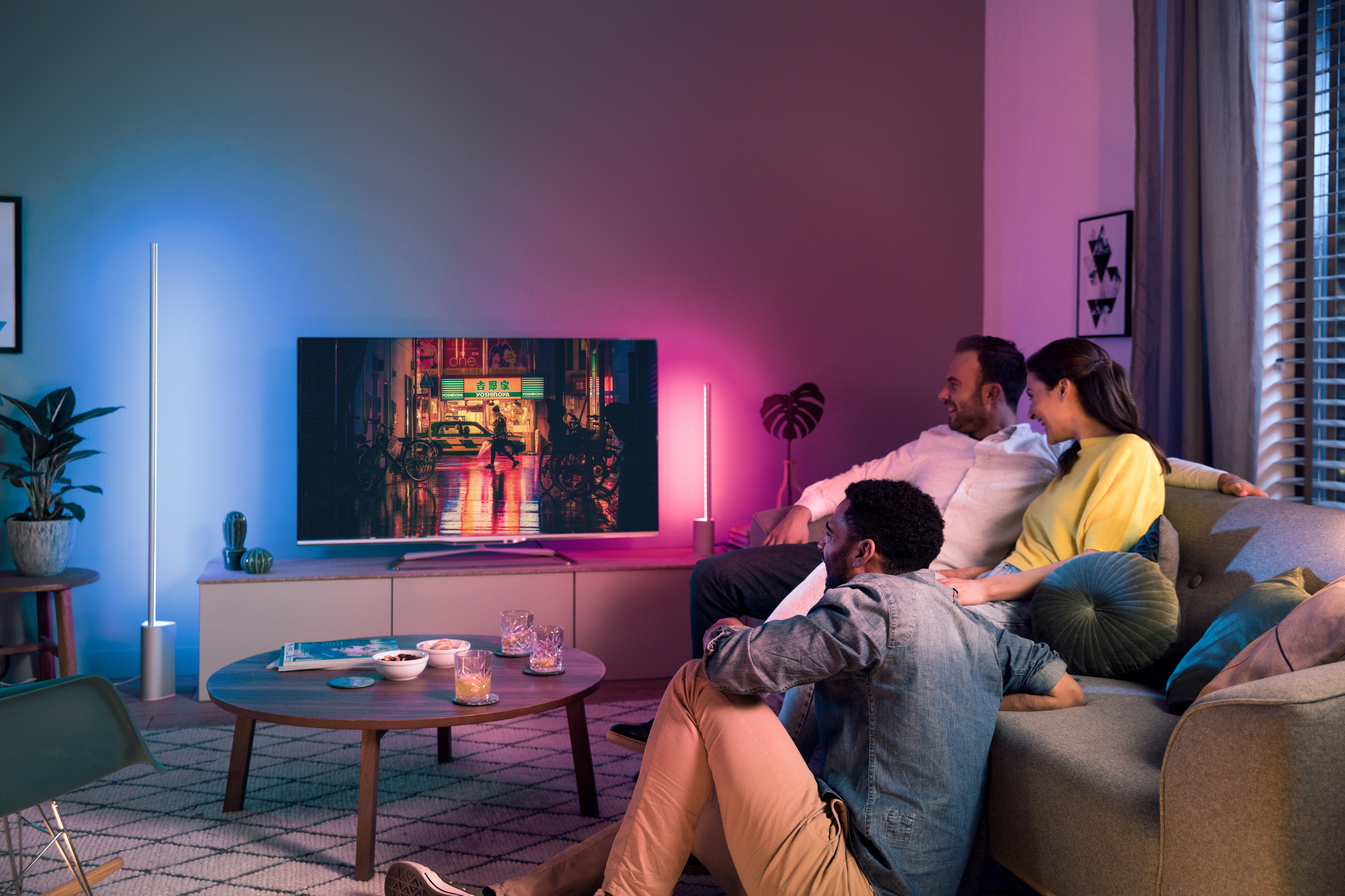 Drei Personen auf einem Sofa schauen fern in einem Raum mit farbigen Lichtern; Couchtisch mit Snacks.