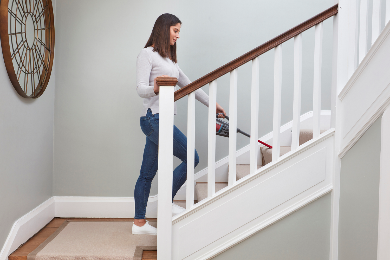 Eine Frau saugt Treppen. Sie ist auf der Treppe und saugt den Teppich. Eine runde Uhr hängt an der Wand.