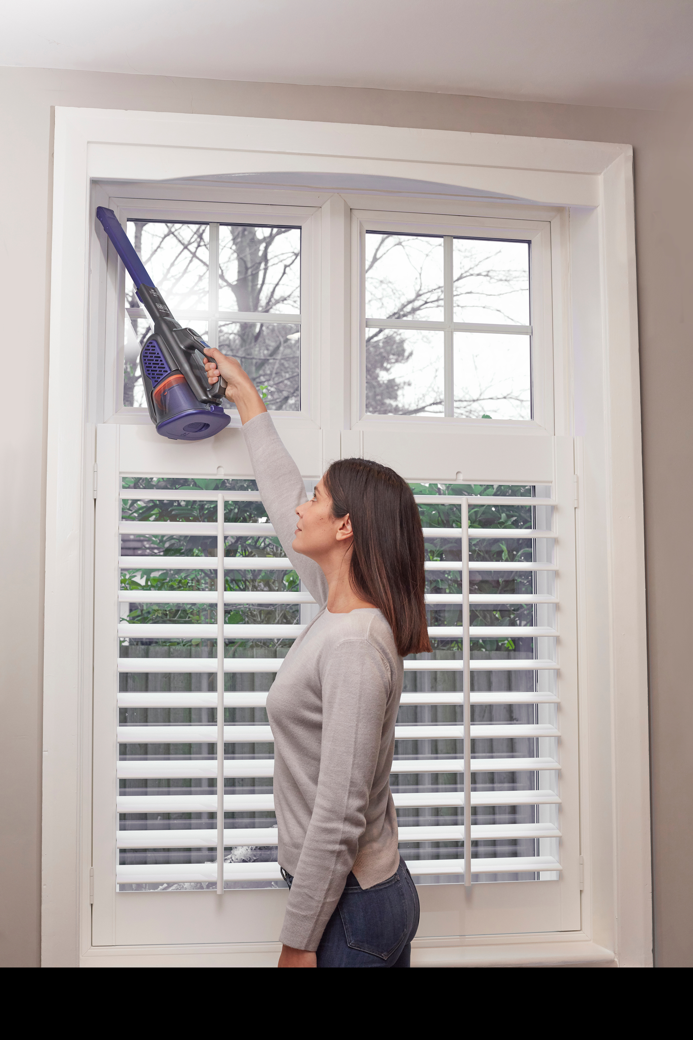 Une femme passe l'aspirateur sur une fenêtre avec un aspirateur à main, près de volets blancs.