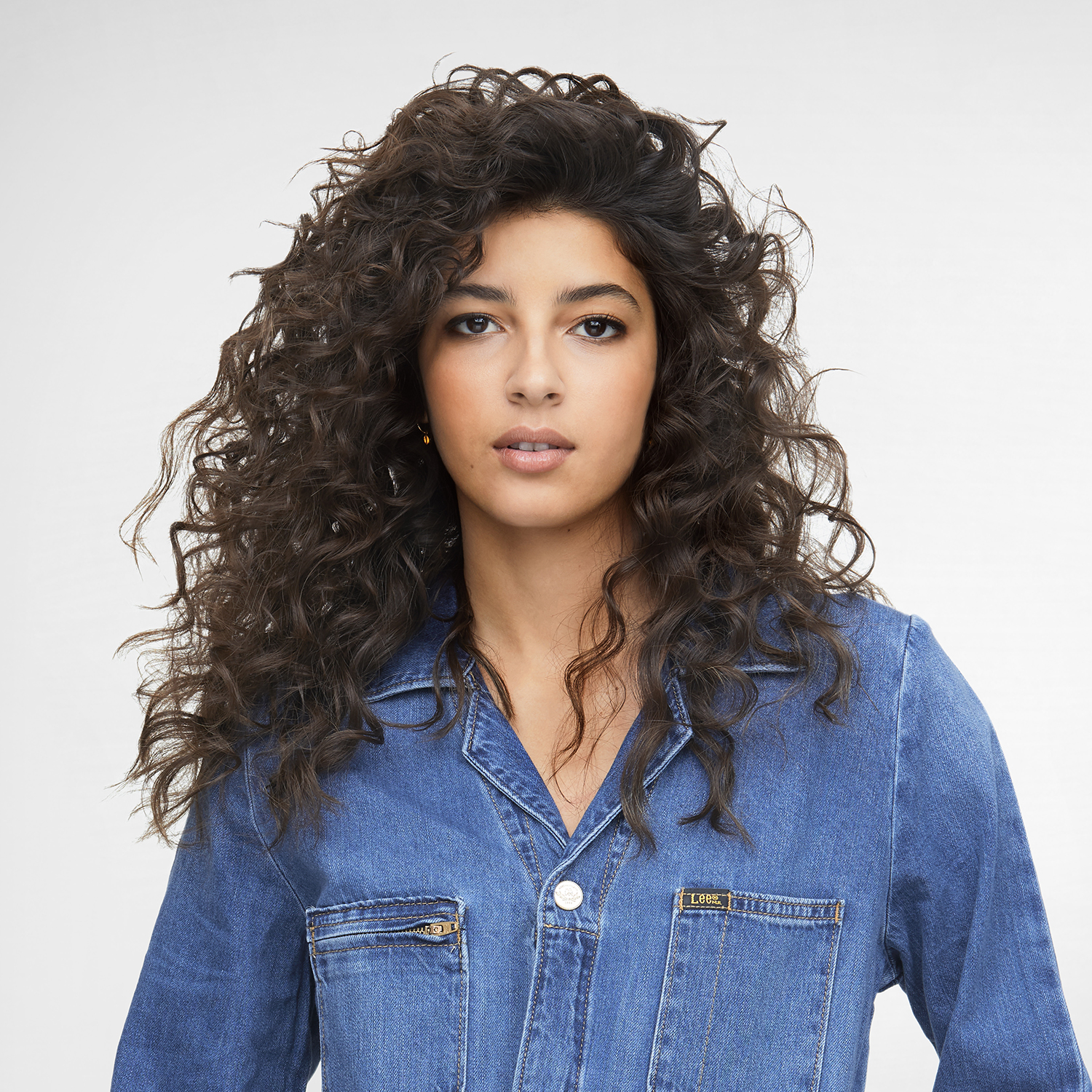 A woman with curly dark hair, wearing a denim shirt, looking towards the camera.