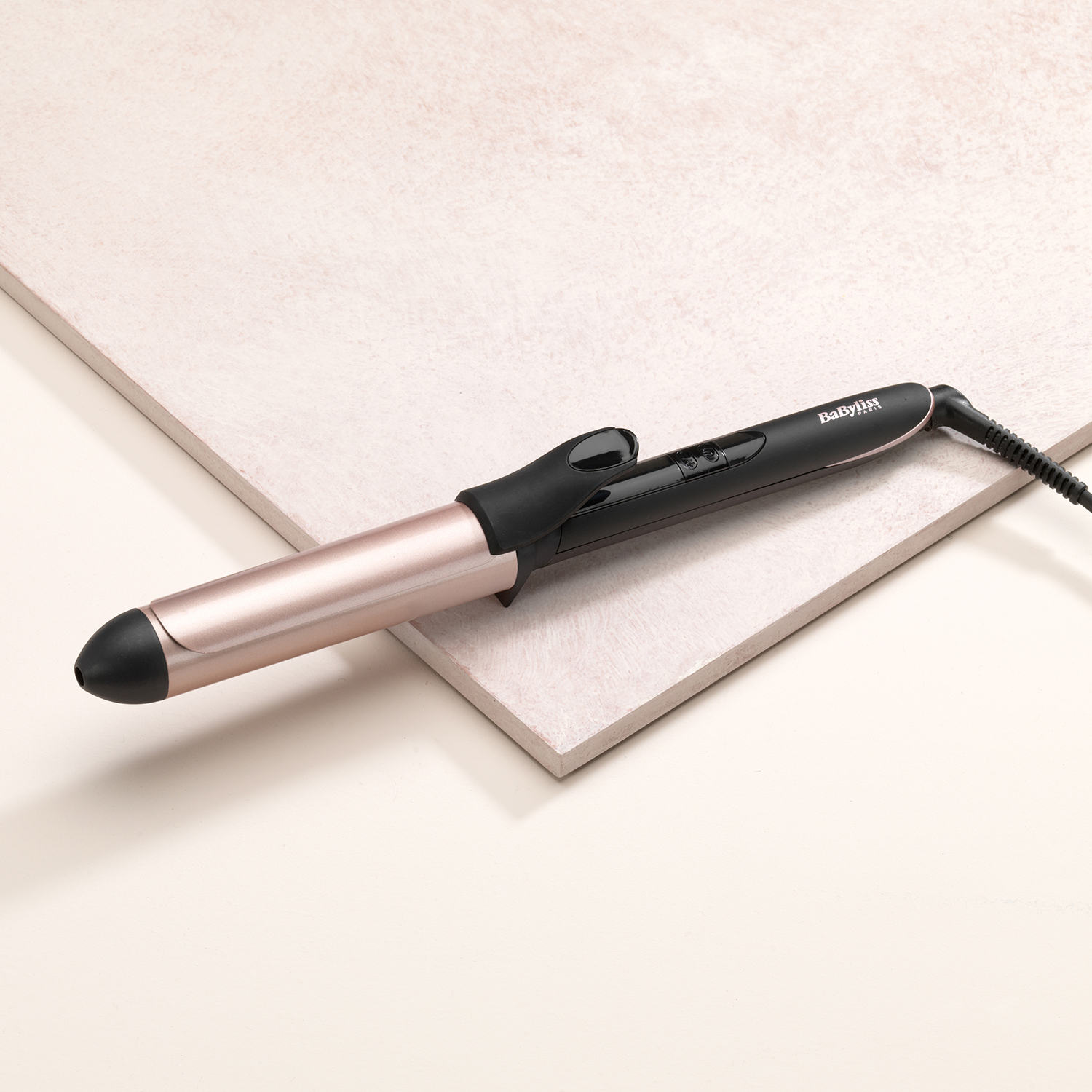 Hair curling iron with a black handle and rose gold barrel. Placed on a pink surface.