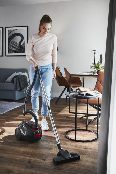 Frau saugt einen Holzboden in einem Wohnzimmer. Ein schwarzer Staubsauger steht auf dem Boden.