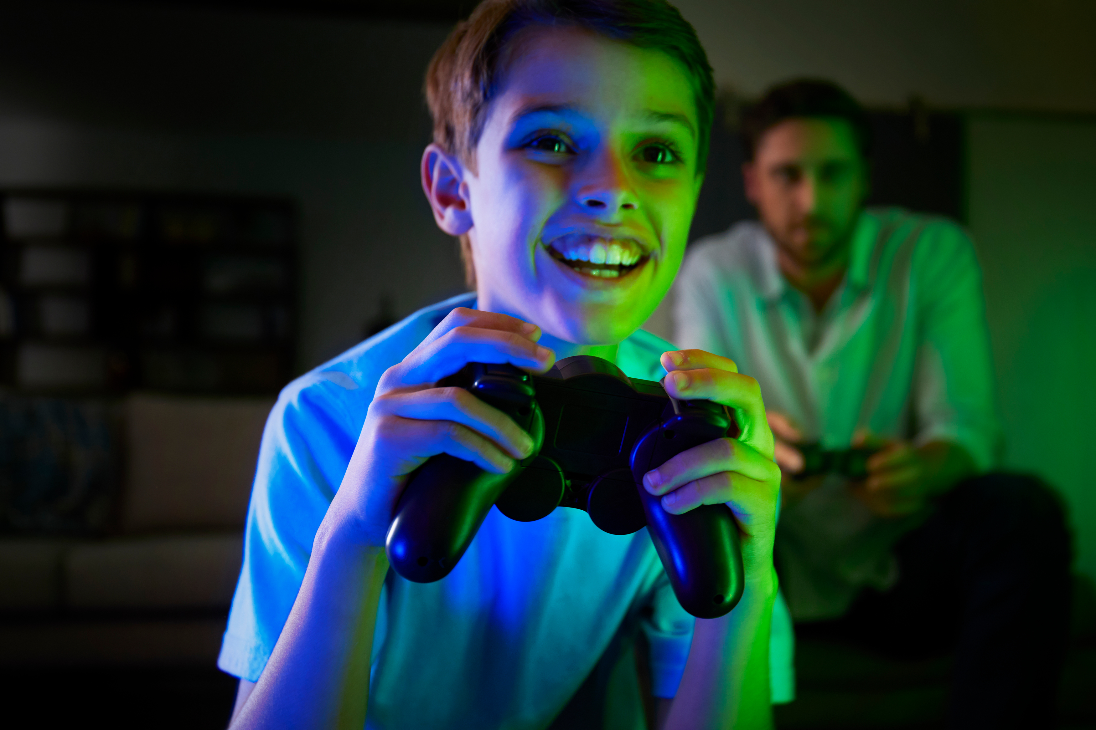 Junge spielt Videospiel mit Controller, lächelt. Vater im Hintergrund spielt auch.