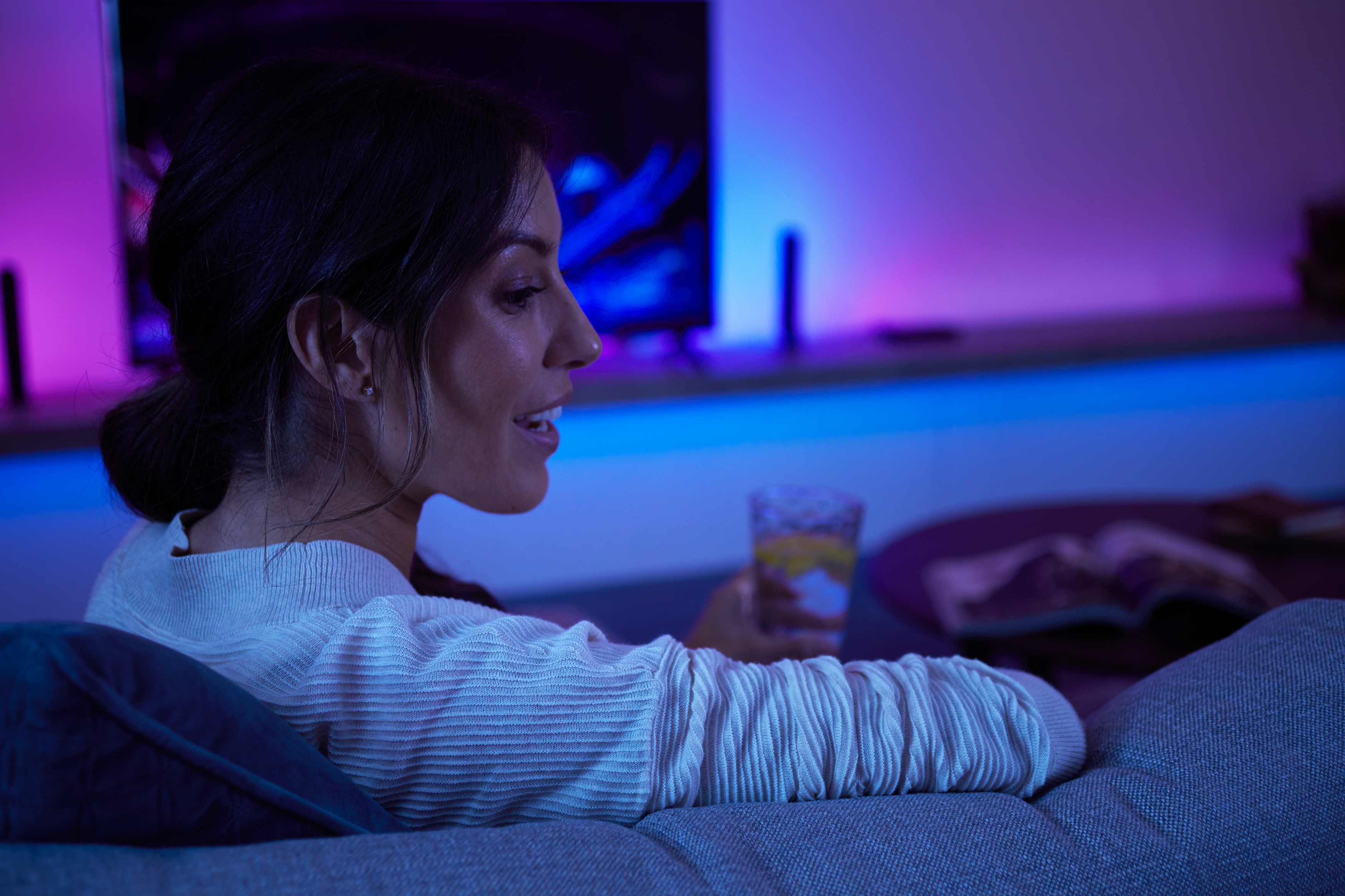 Eine Frau sitzt auf einem Sofa und hält ein Glas. Im Hintergrund sind bunte Lichter zu sehen. Auf dem Tisch liegt ein Buch.