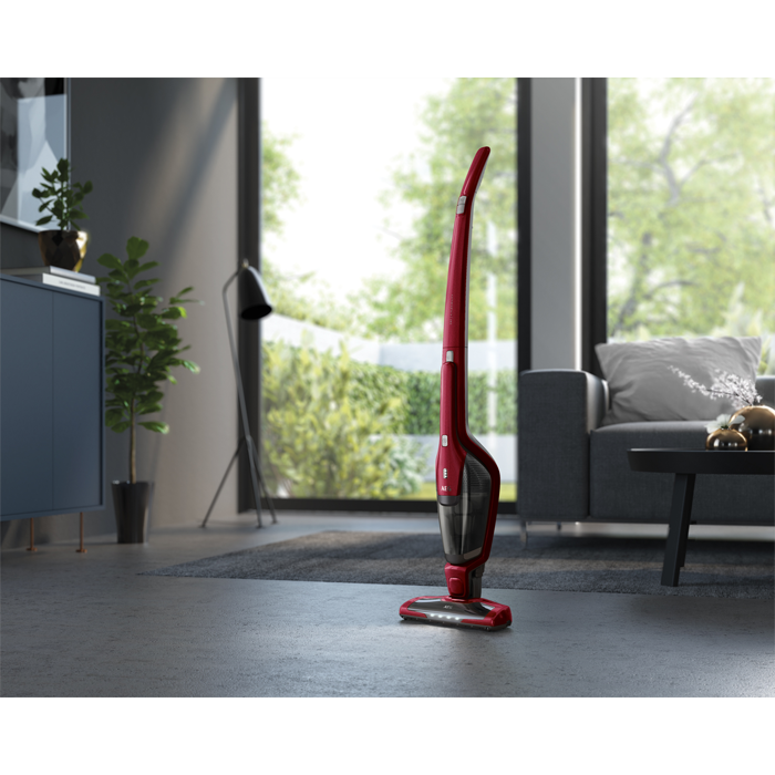 Ein roter kabelloser Staubsauger steht in einem modernen Wohnzimmer. Im Hintergrund sind ein Sofa und Pflanzen zu sehen.
