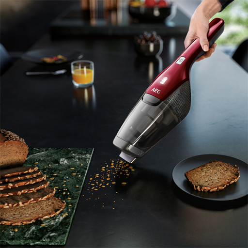Ein rot-schwarzer Handstaubsauger, der Krümel von einem schwarzen Tisch saugt. Brot, Saft und Obst sind im Hintergrund.