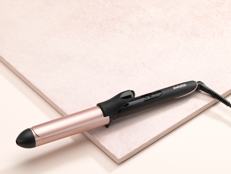 Un fer à friser avec une poignée noire et un canon doré, posé sur une surface rose et beige.