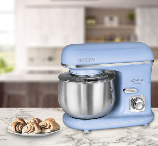 Ein blauer Küchenmixer mit Edelstahlschüssel. Zimtschnecken befinden sich auf einem Teller links.