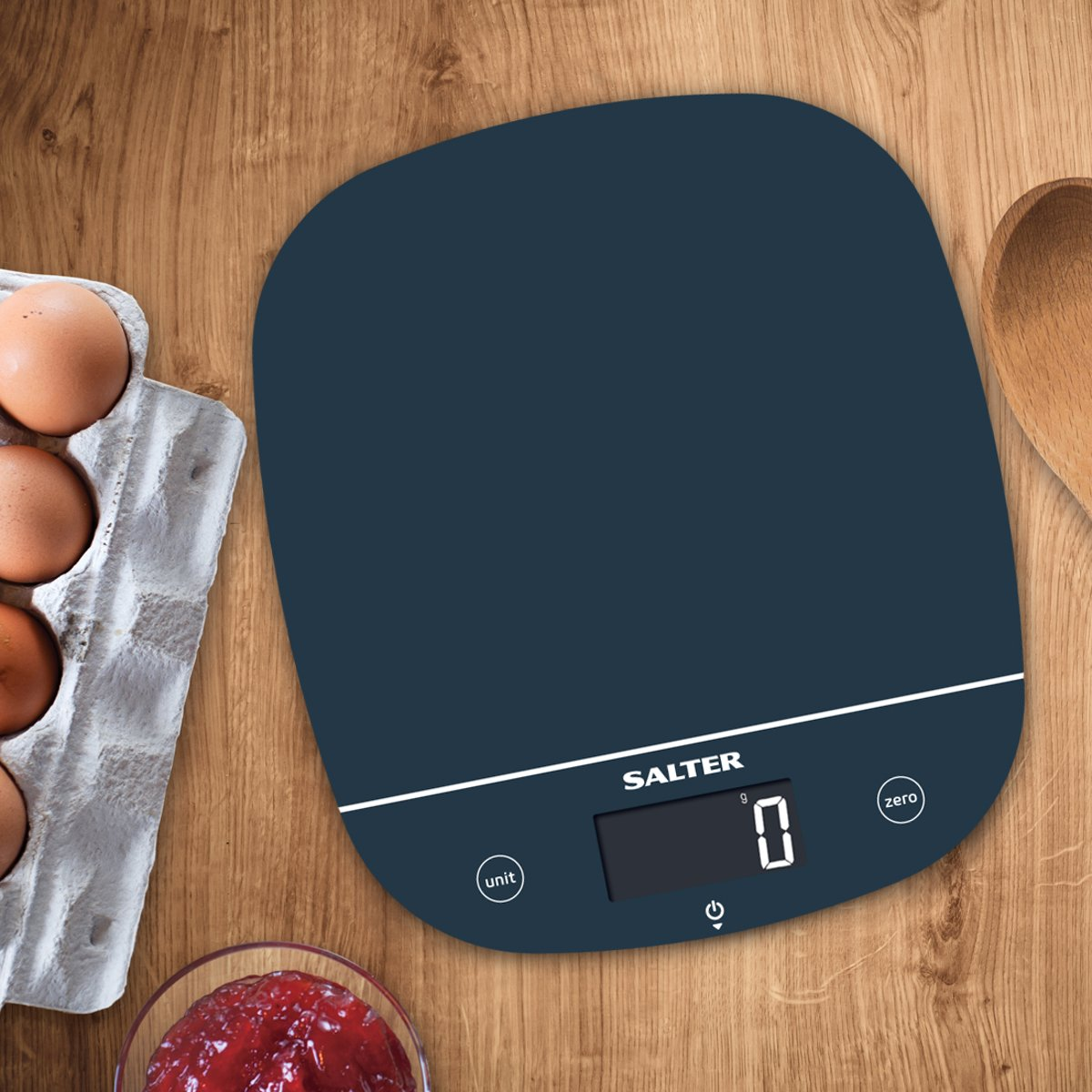 Blue digital kitchen scale with eggs, jam, and a wooden spoon.