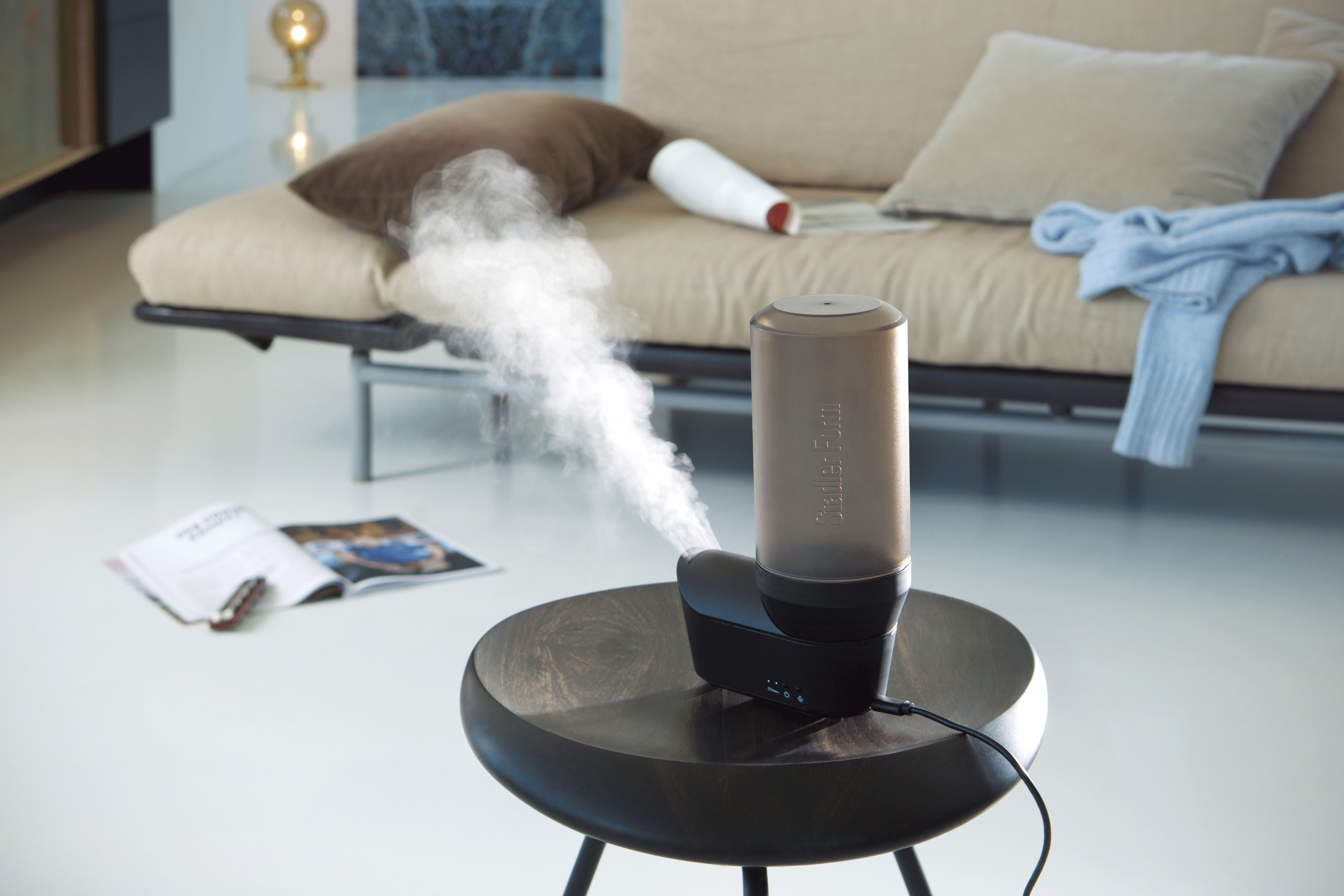 Un humidificateur d'air noir et marron avec de la vapeur qui sort, sur une table. Il y a un canapé derrière.