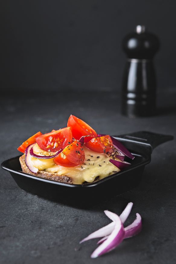 A small raclette dish with tomatoes, onions, and cheese on a dark surface.