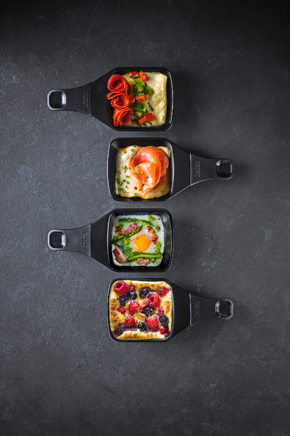 Four small black pans with food. They contain different dishes: a berry dessert, eggs, and meat.