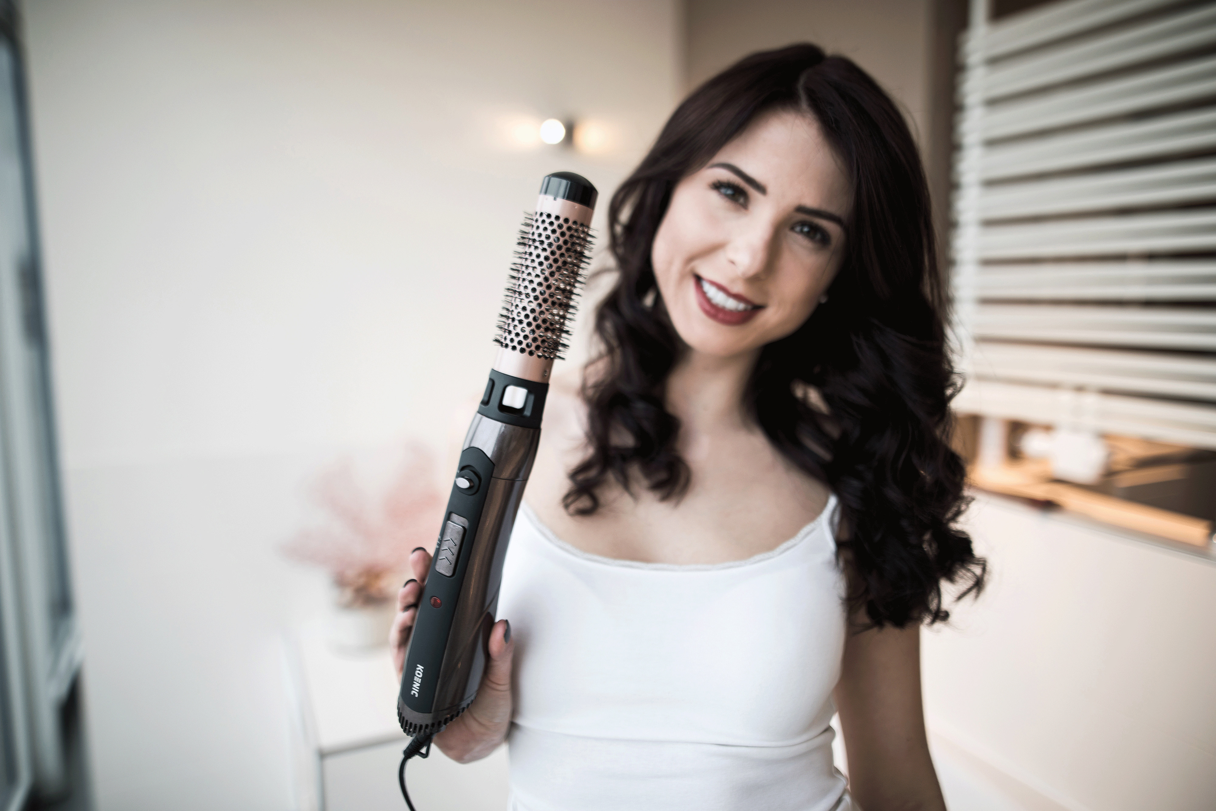 Eine Frau hält einen Haarstyler und lächelt in einem hellen Raum in die Kamera.