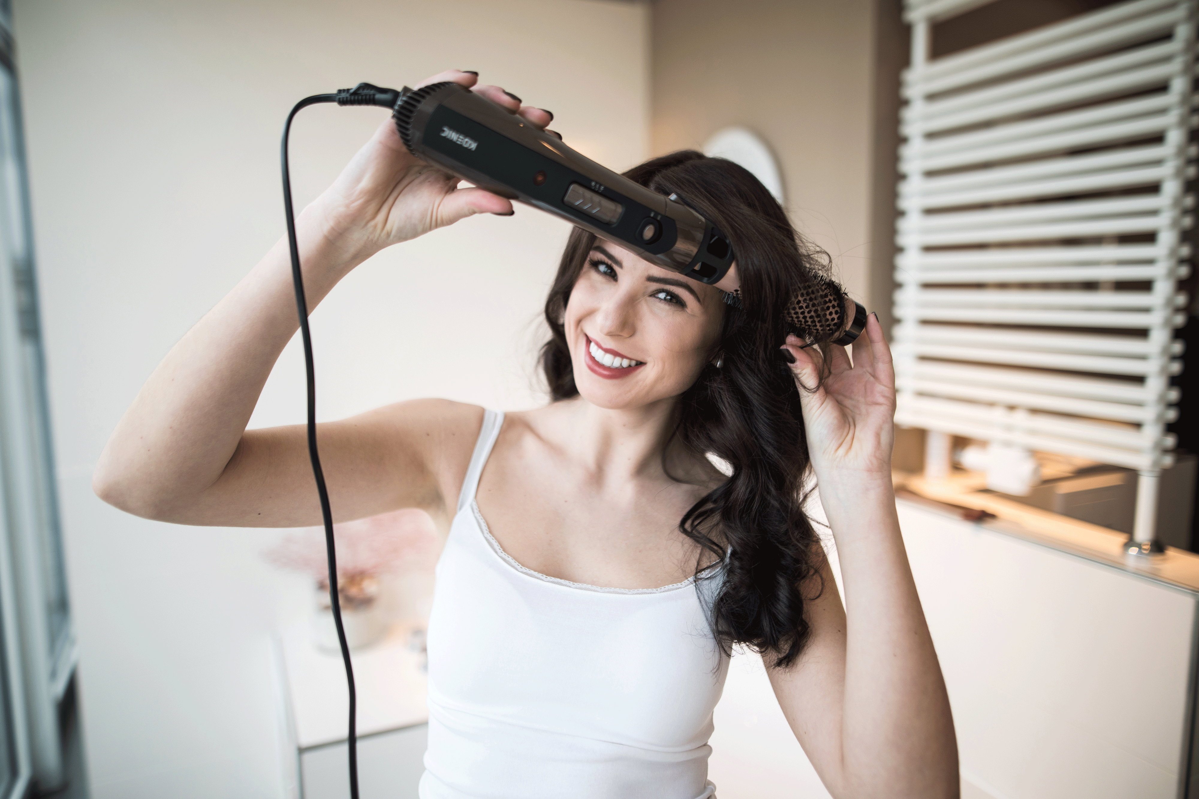 Frau stylt Haare mit Lockenstab. Lächelt, weißes Oberteil, Spiegel im Hintergrund.