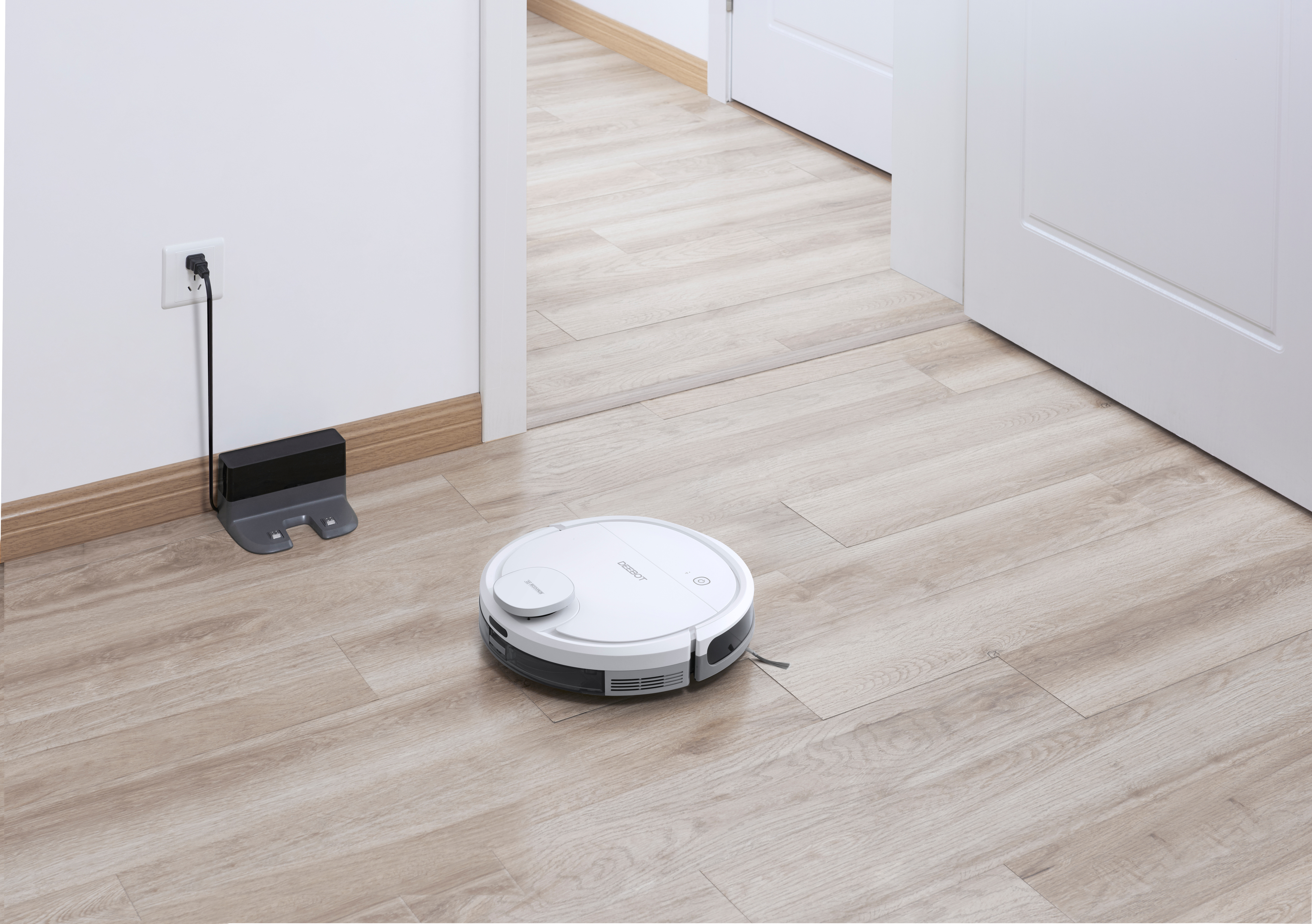 Weißer Roboterstaubsauger auf Holzboden neben Ladestation und offener Tür.