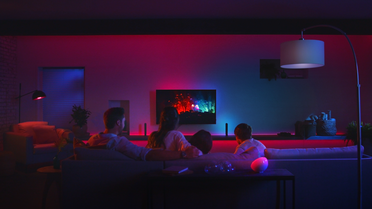 Eine Familie schaut fern in einem Wohnzimmer, das von bunten Lichtern beleuchtet wird. Eine Stehlampe steht in der Ecke.