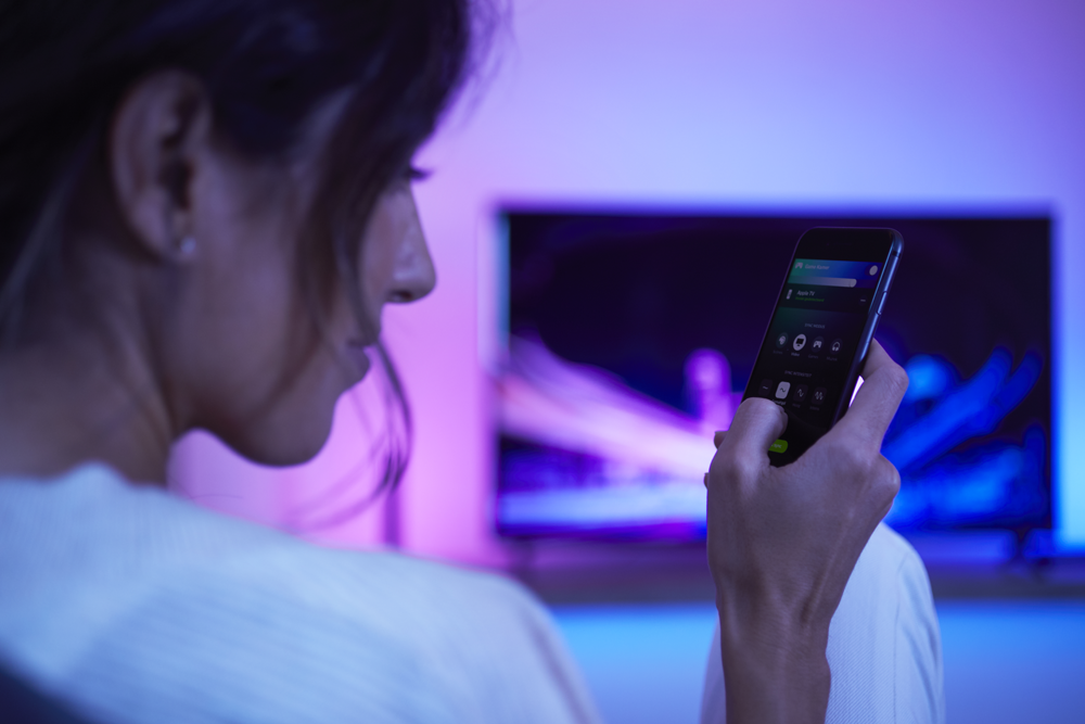 Frau steuert Smart-Home-Beleuchtung auf ihrem Telefon, Fernseher im Hintergrund.