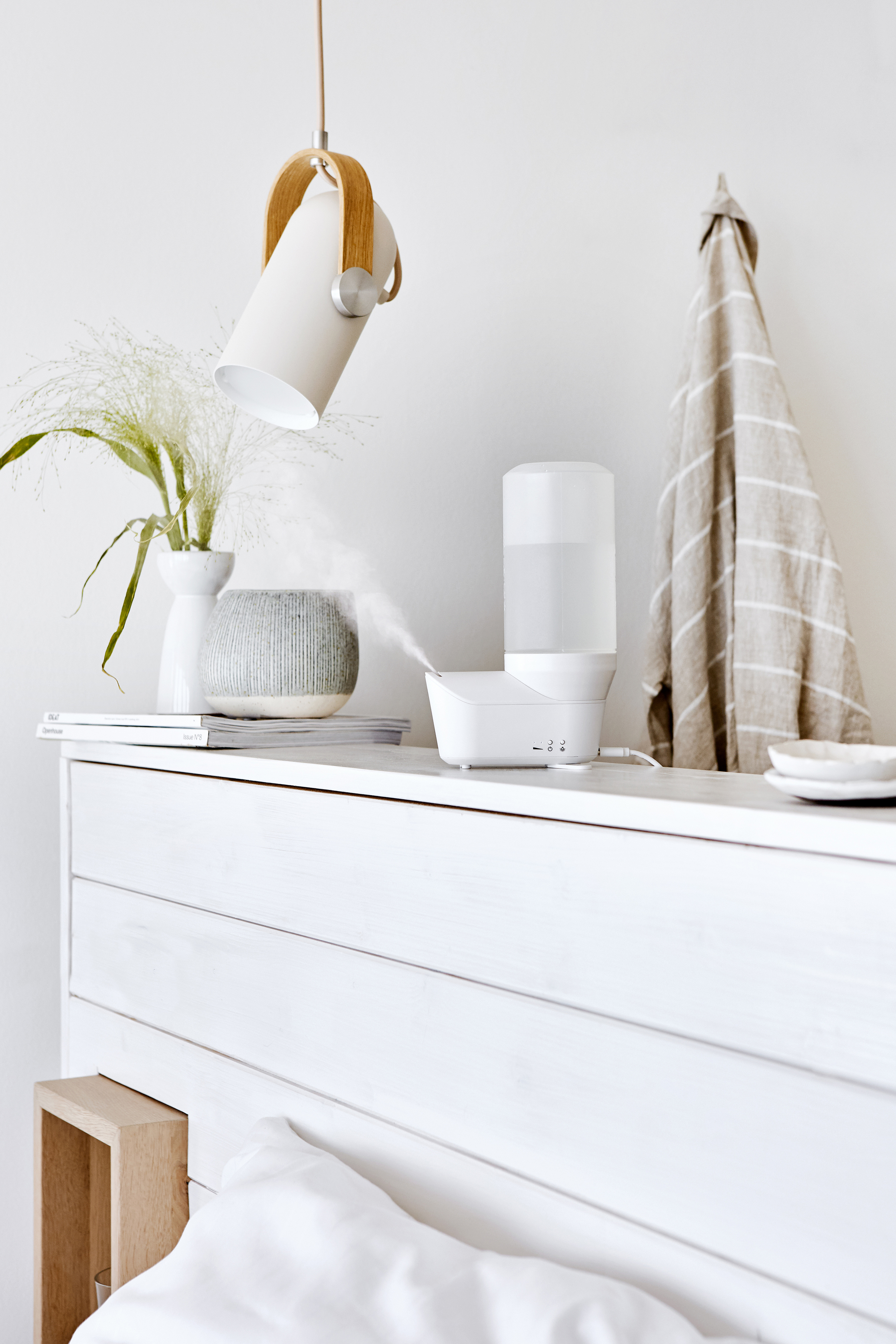 Weißes Schlafzimmer mit Lampe, Vase mit Grünzeug, Luftbefeuchter und Handtuch an weißer Wand.
