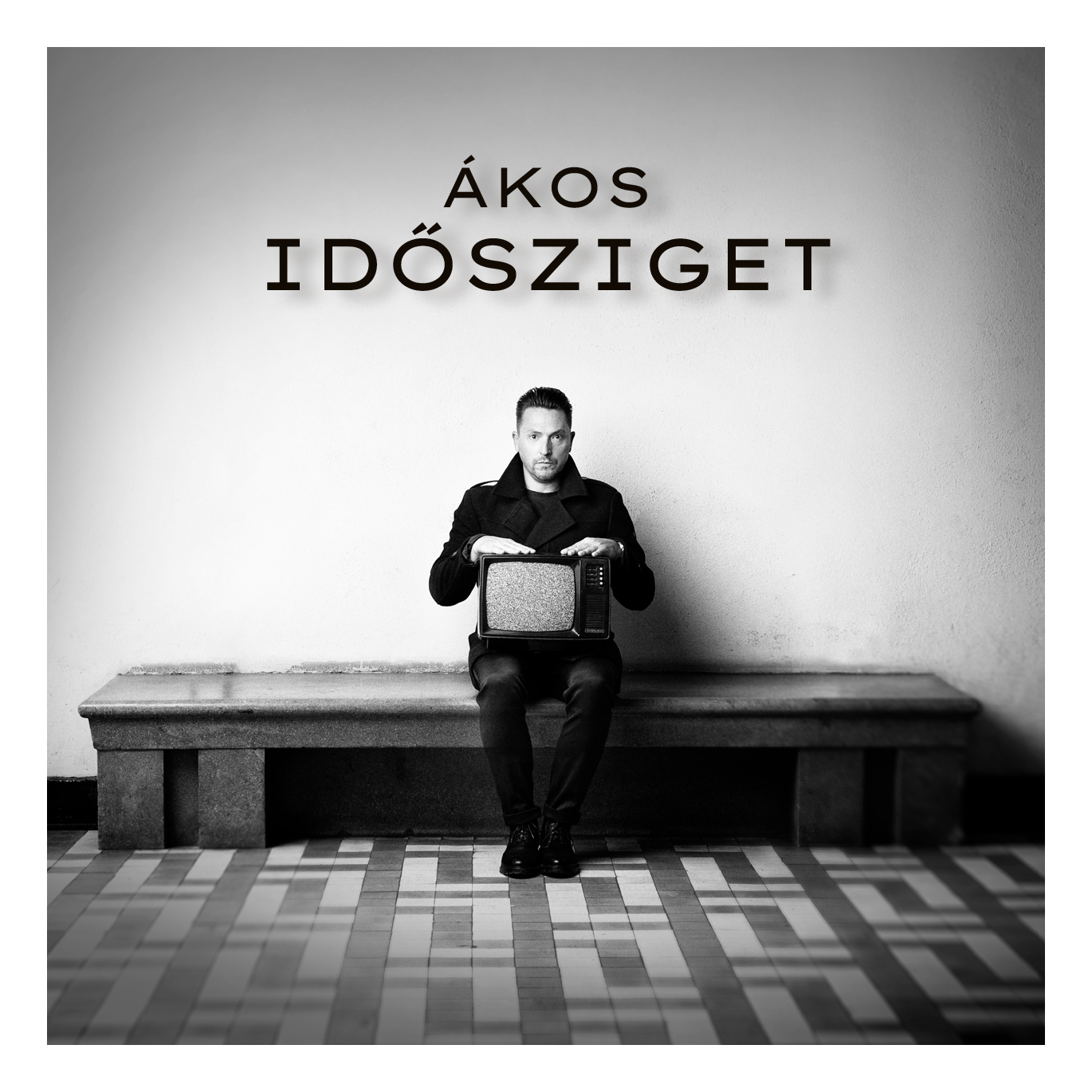 Egy szürkeárnyalatos kép egy férfiról, aki egy televíziót tart. A 'Ákos' és 'Idősziget' szavak felette.