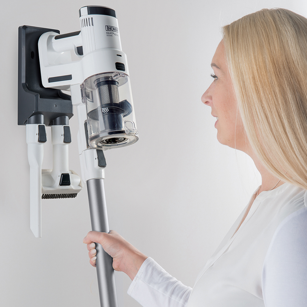 Blonde Frau hält einen weißen und grauen Staubsauger neben einem wandmontierten Ladegerät.