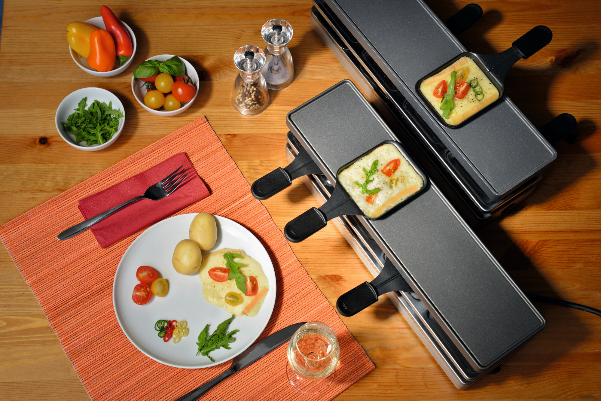 Impostazione della cena raclette. Due griglie per raclette con formaggio, verdure, un piatto con contorni e un bicchiere di vino.
