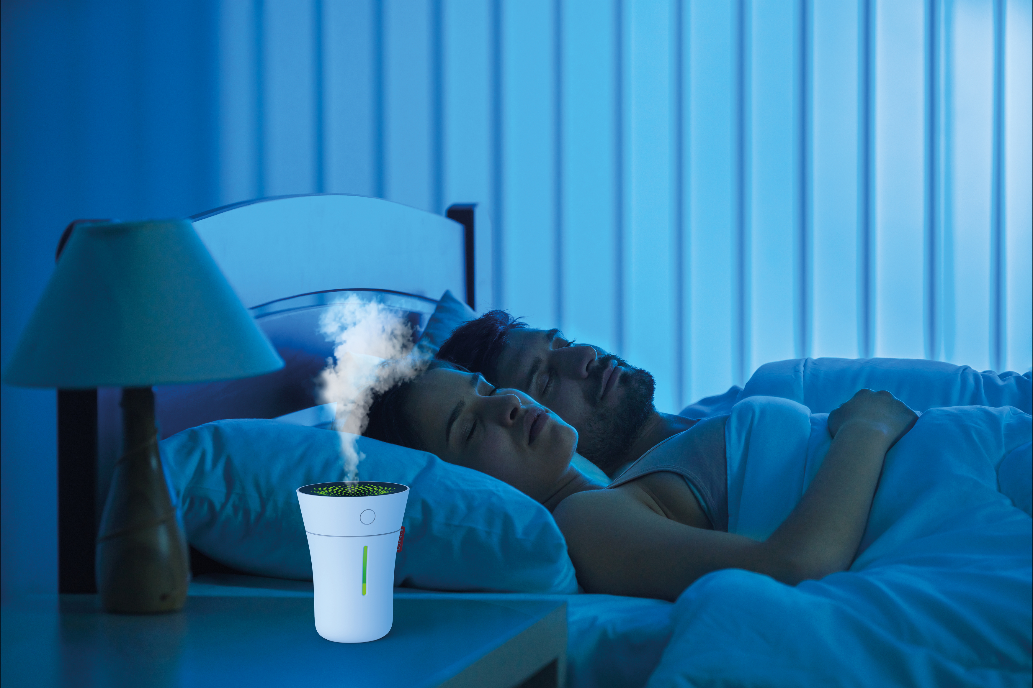 Ein Paar schläft im Bett, ein Luftbefeuchter auf dem Nachttisch gibt Dampf ab.