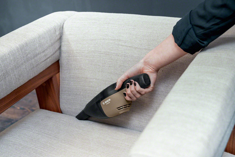 Hand saugt ein helles Sofa mit einem tragbaren Staubsauger.